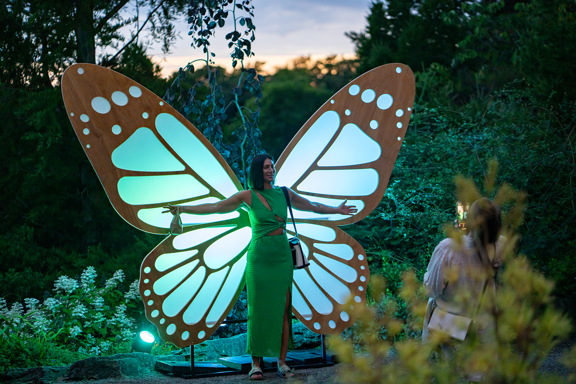 Woman posing with large, light up butterfly wings in the garden at night