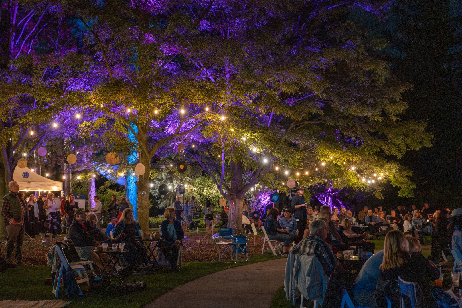 evening event in the summer with visitors seated watching a performance, trees lit with string lights and up lighting at Rock Garden