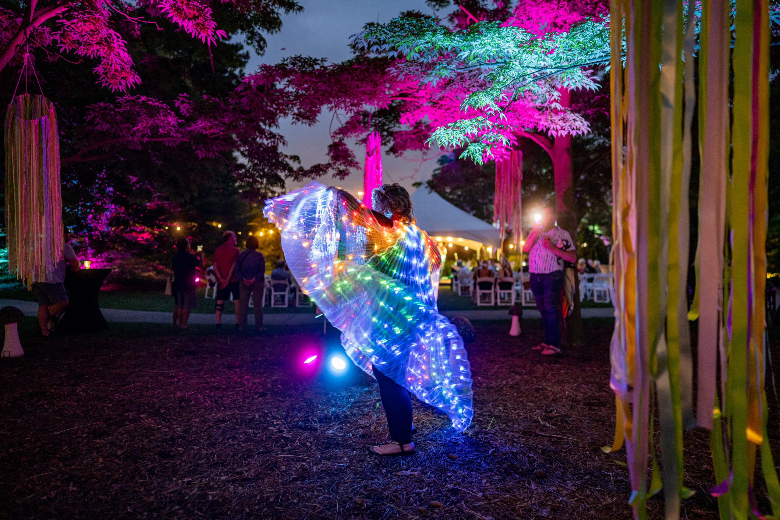 A guest dances under illuminated trees at an outdoor nighttime event, wearing glowing LED wings that shimmer in rainbow colors.