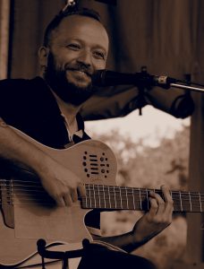 Juneyt sitting on a stool on a stage playing guitar