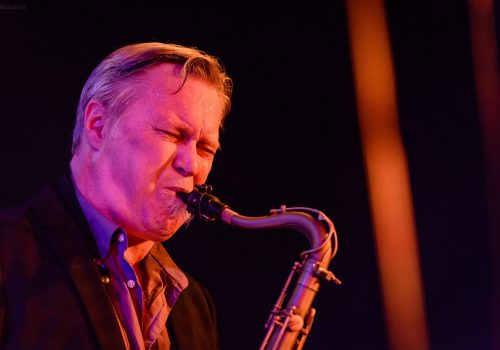 kirk_macdonald_head_shot_color_2 Kirk MacDonald playing saxophone in a dark, dramatically lit space, wearing a suit