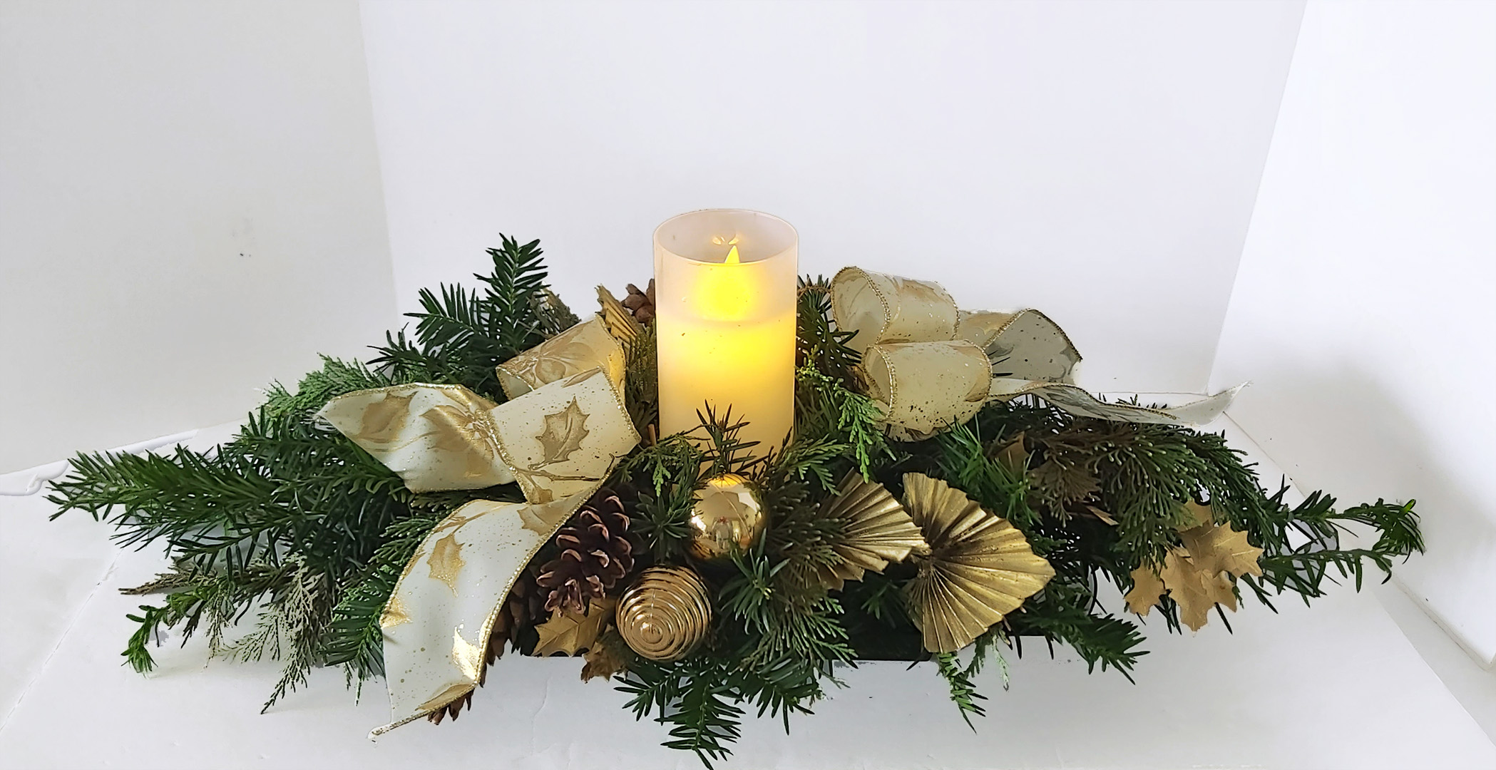 Evergreen table centrepiece featuring an everlasting candle nestled among the fresh scent of evergreens, complimented with classic gold accents, pinecones and finished with the delicate sparkle of ribbon